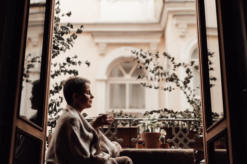 A woman sitting on a balcony with eyes closed holding a mug and meditating.