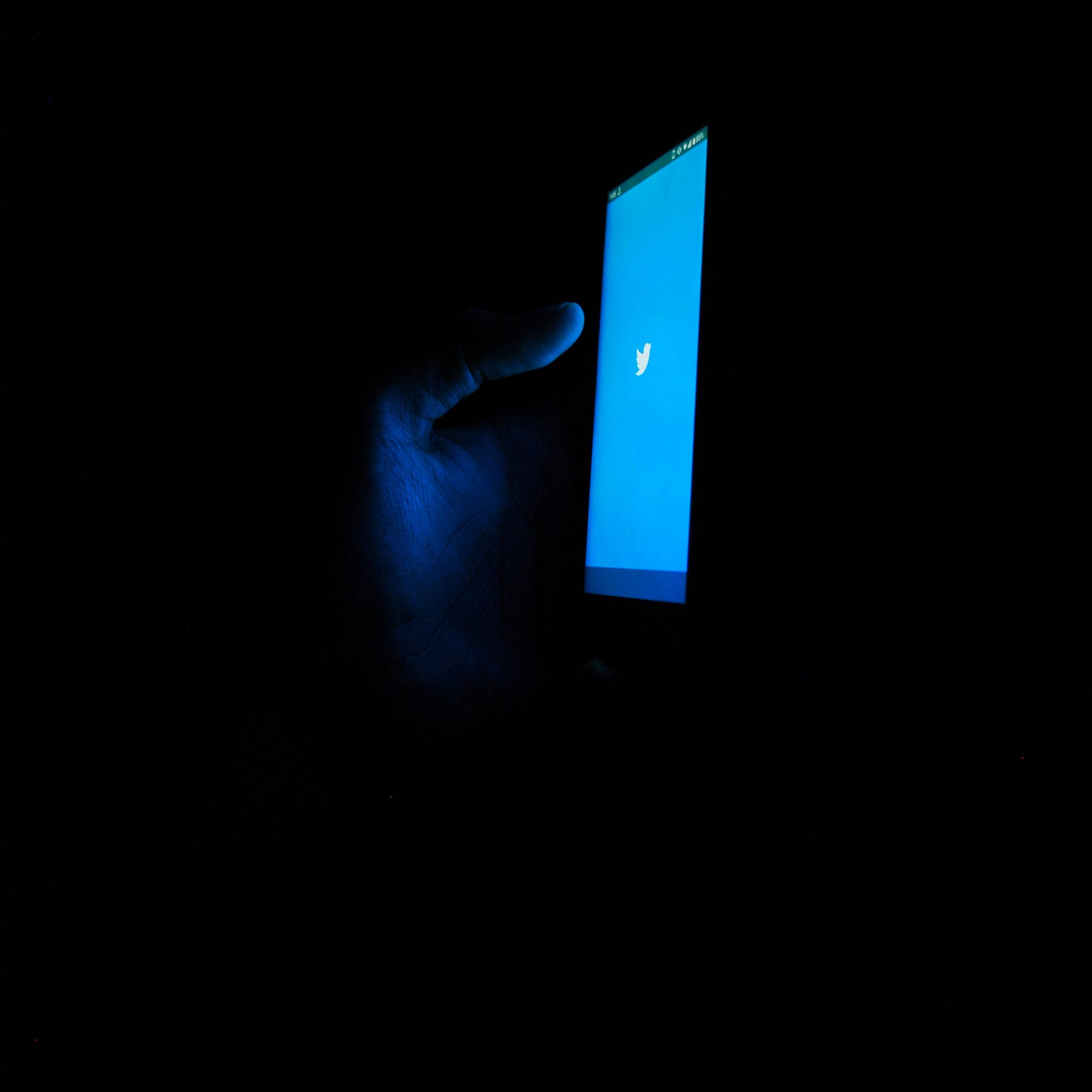 A hand holding a phone in the dark, it has a blue screen with the twitter logo.
