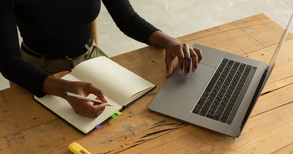 Person's hands working at a laptop with a pen and notebook on the desk.