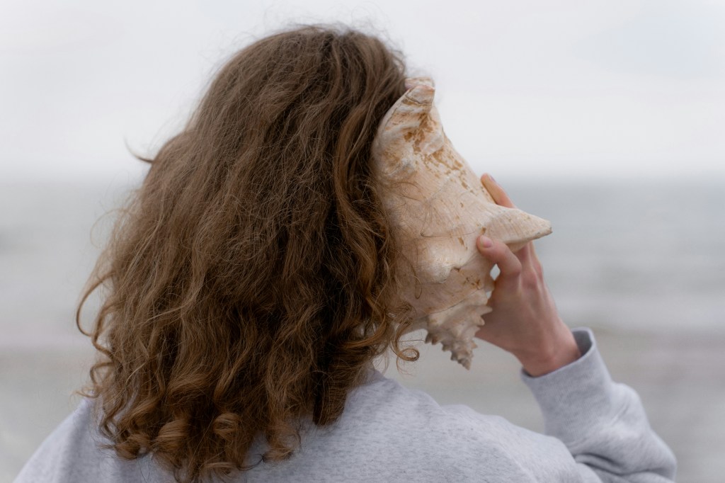 Woman standing in front of the ocean, holding a large shell to her ear and listening.