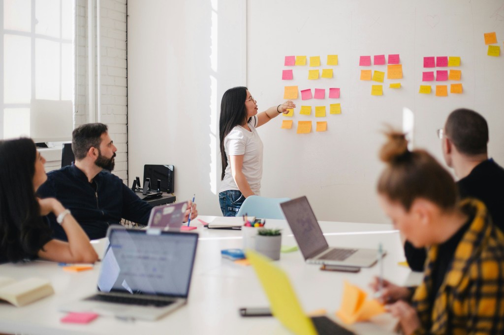 A team works around a table with laptops and a woman stands at the wall with sticky notes to plan their work schedule.