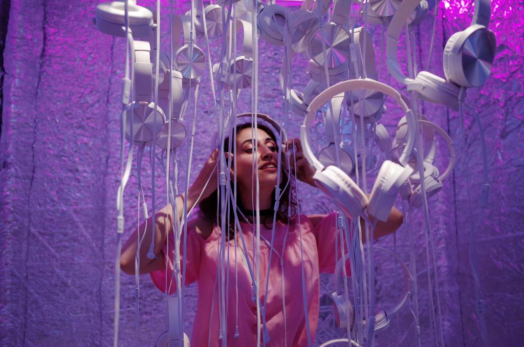 A woman standing in a shimmery, purple room surrounded by silver headphones dangling from the ceiling.
