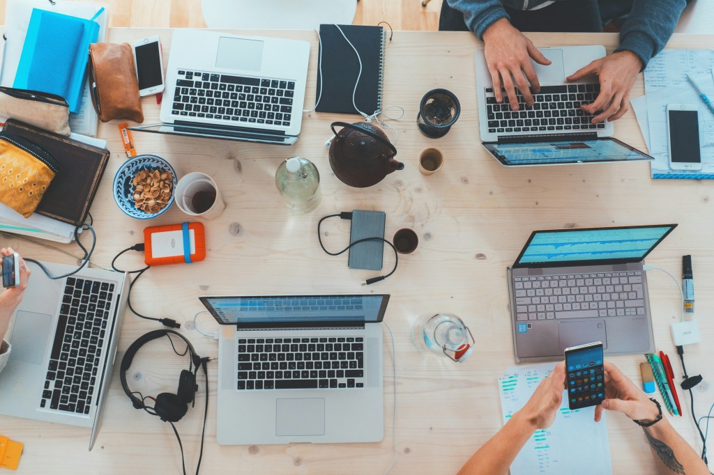 Bird's eye view of a work table with laptops, phones tea and snacks as people work together.